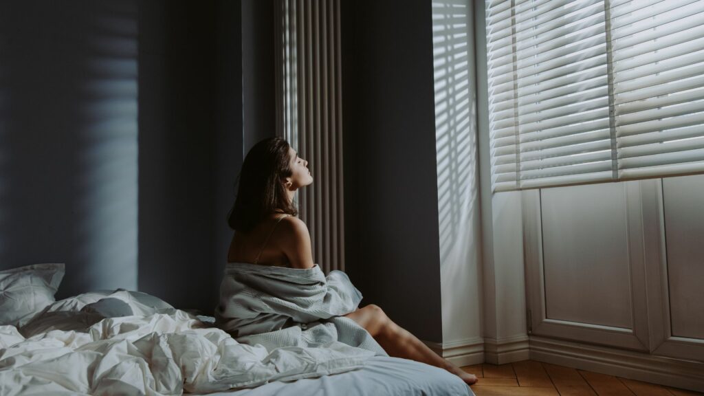 Eine Frau sitzt morgens auf ihrem Bett und blickt aus dem Fenster.